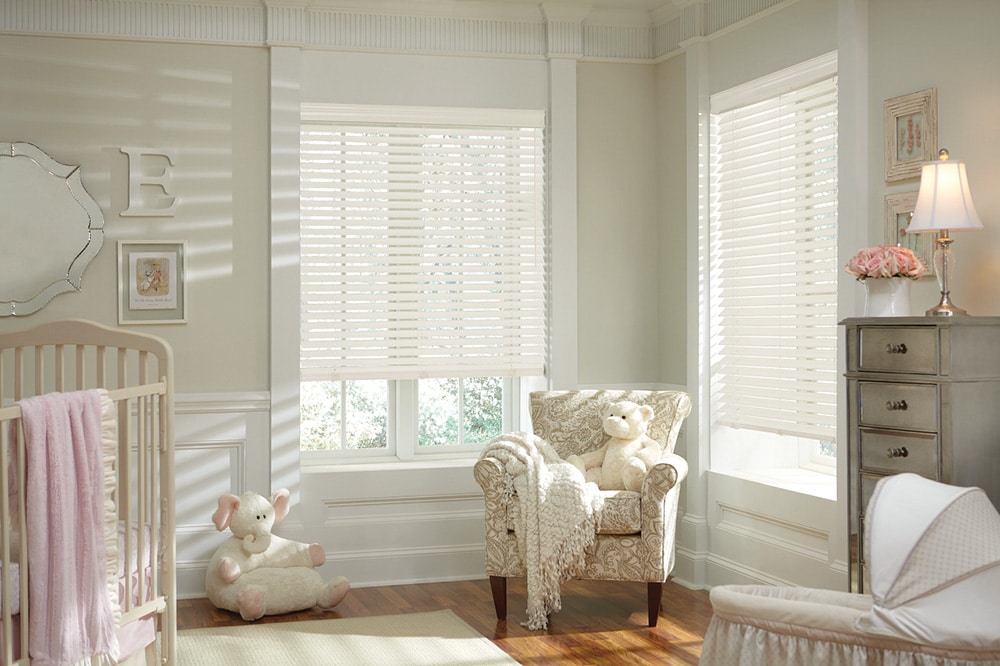A soft neutral nursery with white wood blinds on two windows.